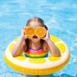 Child floating in a pool on an inflatable ring while holding orange slices over their eyes